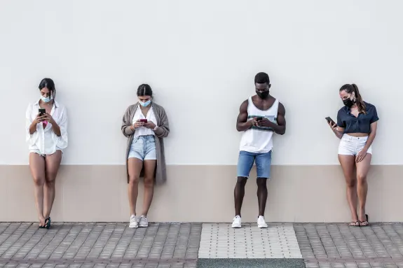 Four people wearing masks standing apart using smartphones against a wall.