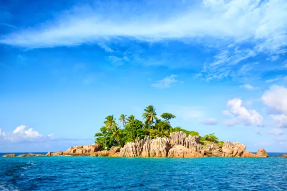 Small tropical island with palm trees and rocks in a bright blue ocean under a clear sky.