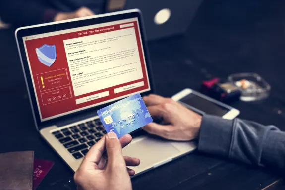 Person holding credit card near laptop displaying ransomware screen, symbolizing online payment fraud.